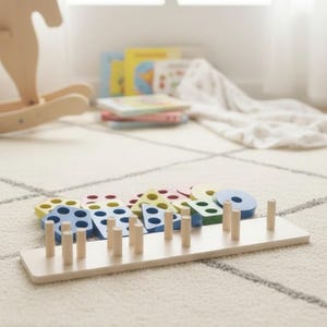 Op de afbeelding: Houten vormsorteerspeelgoed met kleurrijke geometrische vormen, waaronder cirkels, driehoeken en vierkanten. De vormen zijn geschilderd in verschillende kleuren zoals blauw, geel en groen. Het speelgoed heeft houten pinnen waarop de vormen passen. De basis is lichtgekleurd hout.