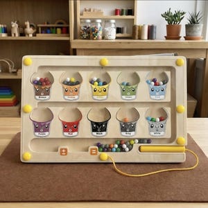 May include: Wooden educational toy featuring buckets labelled with colours and filled with matching balls. The toy has a maze design with a yellow handle and buttons. The background includes wooden shelves and plants.