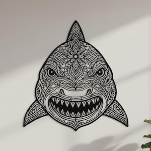 May include: Black metal shark wall art with intricate floral and geometric patterns. The shark's open mouth reveals sharp teeth. The artwork is mounted on a white wall, adding a striking contrast. A unique home decor piece.
