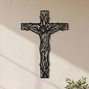 Puede incluir: Arte mural de metal negro que representa a Jesús en la cruz. La cruz y la figura están estilizadas con un diseño similar a un árbol. La obra de arte está montada en una pared de color claro. La imagen también muestra una mesa de madera con velas, un libro y un jarrón con vegetación.