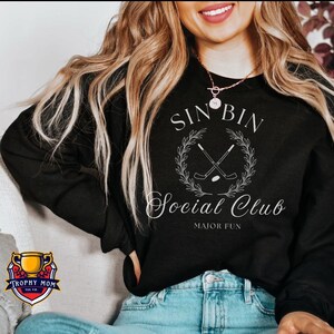 May include: Black sweatshirt with the text "SIN BIN Social Club MAJOR FUN" in white script. The design includes crossed hockey sticks within a laurel wreath. The sweatshirt is worn with blue jeans.