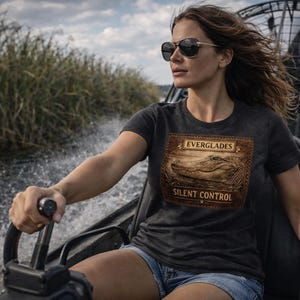 May include: Dark gray t-shirt with a brown graphic featuring an alligator and the words "Everglades" and "Silent Control." The shirt is worn by a person on an airboat, with water and tall grass in the background.