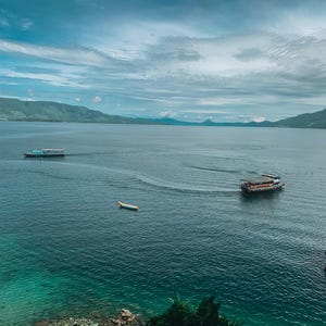 以下が含まれることがあります： ボートと山々を背景にした湖の美しい景色。水は深い青緑色で、空は曇りです。小さな黄色いボートや2隻の大きな旅客フェリーなど、水上にはいくつかのボートが見えます。