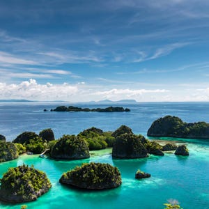 May include: Aerial view of a tropical seascape with turquoise water and small islands covered in lush green vegetation. The sky is a vibrant blue with scattered clouds, creating a picturesque landscape.