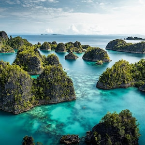 May include: Aerial view of a tropical seascape featuring numerous small, rocky islands covered in lush green vegetation. The islands are surrounded by clear, turquoise water under a partly cloudy sky, creating a vibrant and scenic landscape.