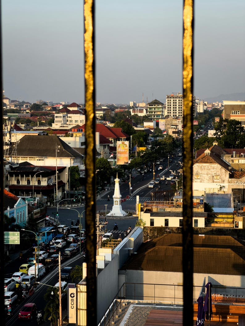 May include: An aerial view of a city street scene, framed by vertical gold-colored bars. The image captures a busy road with cars, buildings, and a white monument in the center. The sky is a soft, muted color.
