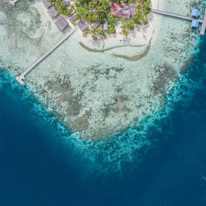 May include: Aerial view of a tropical island with a village, surrounded by turquoise water and a deep blue ocean. The island features white sand beaches, palm trees, and buildings with blue and red roofs. Wooden piers extend into the clear water.