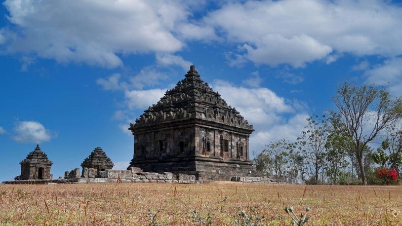May include: A landscape view of the Ratu Boko temple complex in Indonesia. The ancient stone structures are set against a bright blue sky with fluffy white clouds. The foreground features dry, golden grass.