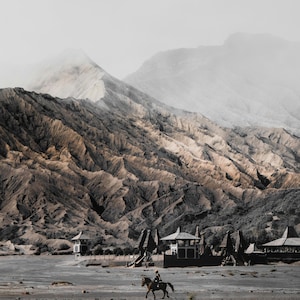 Puede incluir: Fotografía de paisaje con una persona montando a caballo a través de una llanura arenosa. En el fondo, montañas y estructuras con arquitectura tradicional. La escena se captura en tonos apagados, centrándose en el entorno natural.