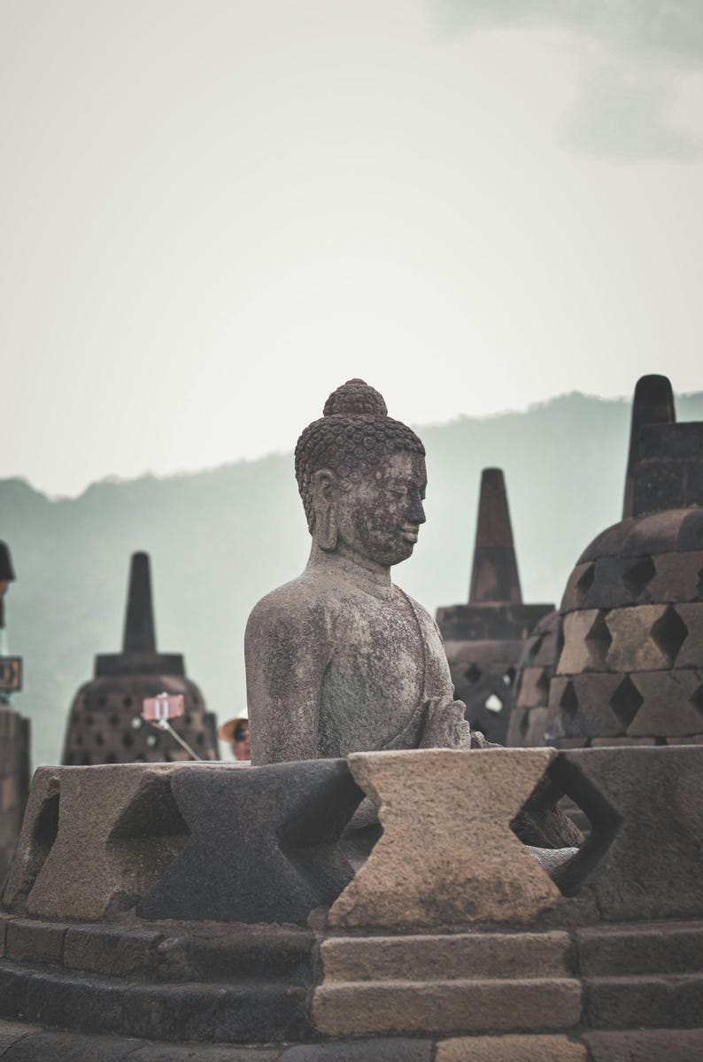 May include: A weathered stone statue of Buddha sits within a stone structure, surrounded by other stupas. The statue is gray and has a serene expression. The background features a mountain range under a cloudy sky.