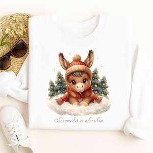 Peut inclure: Sweat-shirt blanc avec un âne de dessin animé portant un bonnet et une écharpe rouges et oranges, assis sur un nuage avec des sapins de Noël. Le texte "Oh, come let us adore him" est imprimé en dessous.