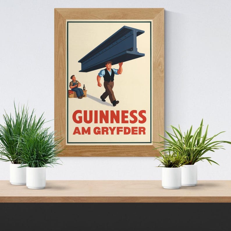 Op de afbeelding: Een ingelijste print met een vintage Guinness-advertentie. De afbeelding toont een man die een grote stalen balk draagt, met een andere man die ernaast zit. De tekst "GUINNESS AM GRYFDER" staat in het rood. De print zit in een houten lijst.