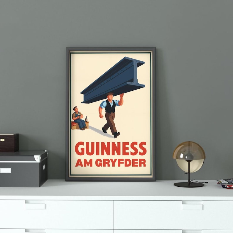 Op de afbeelding: Ingelijste vintage Guinness-advertentie. De poster toont een man die een grote blauwe stalen balk draagt, met een andere man in de buurt. De tekst "GUINNESS AM GRYFDER" wordt in rood en wit weergegeven.