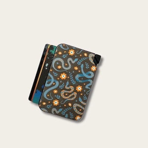 May include: A dark brown wallet with a pattern of blue snakes and orange flowers. The wallet is next to a card holder with a colorful geometric design. The wallet has a black tab on the side.