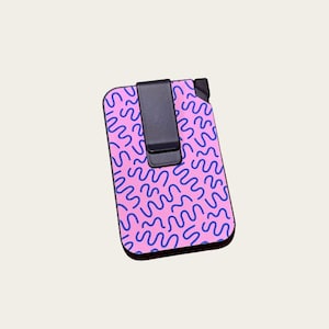 May include: A rectangular card holder with a black clip. The card holder has a pink background with a blue squiggly pattern. The edges of the card holder are black.