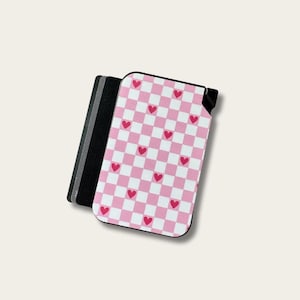 May include: A phone case with a pink and white checkered pattern and red hearts. The case has a black border and is next to a stack of black and gray cards. The design is a cute and playful accessory.