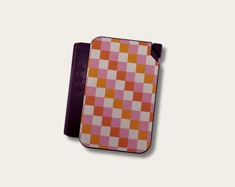 May include: A rectangular lighter with a pink, orange, and white checkered pattern. A dark purple case is next to the lighter. The lighter has a rounded top and a small opening for the flame.