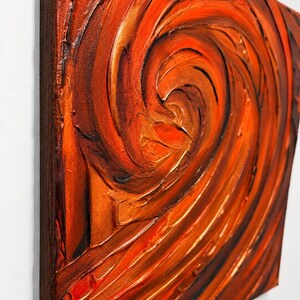 May include: A square canvas painting featuring thick, textured impasto brushstrokes in shades of vibrant orange, deep red, and dark brown. The paint is applied in a swirling, circular motion, creating a dynamic, raised surface.