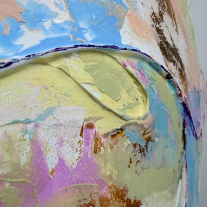 Op de afbeelding: Een abstract schilderij met dikke, getextureerde impasto-lagen van olie- of acrylverf. De compositie toont een palet van pastelblauw, zachtgeel, lichtroze, wit en metallic goud, met een donkerpaarse lijn die een gebogen vorm definieert.