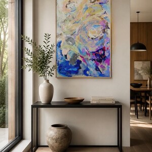 Op de afbeelding: Een rechthoekig abstract schilderij met wervelende penseelstreken in blauw, roze, geel en wit, ingelijst in licht hout. Het kunstwerk hangt boven een zwarte metalen consoletafel met een keramische vaas, een houten schaal en twee boeken.