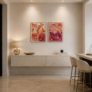 May include: Two rectangular framed abstract paintings featuring vibrant swirls of red, pink, and orange paint. The artwork is displayed side-by-side on a plain wall above a long, wall-mounted white marble cabinet with subtle grey veining.