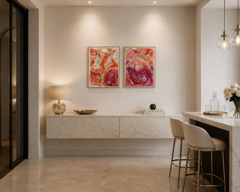 May include: Two rectangular framed abstract paintings featuring vibrant swirls of red, pink, and orange paint. The artwork is displayed side-by-side on a plain wall above a long, wall-mounted white marble cabinet with subtle grey veining.