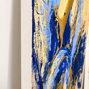 May include: A rectangular canvas painting featuring thick, textured impasto brushstrokes in shades of deep navy blue, light sky blue, and metallic gold. The paint is applied in heavy layers, creating a raised, tactile surface on the canvas.