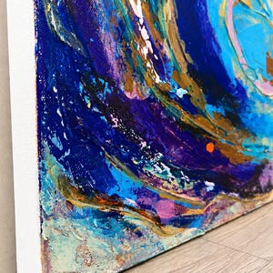 May include: An abstract painting on a rectangular canvas featuring thick, textured layers of acrylic paint in shades of deep navy blue, bright turquoise, metallic gold, and soft pink, with visible brushstrokes and impasto techniques throughout.