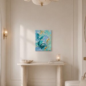 May include: A rectangular canvas painting featuring abstract brushstrokes in shades of teal, light blue, lavender, and white, accented with metallic gold leaf details. The artwork is displayed on a wall above a light-colored console table.