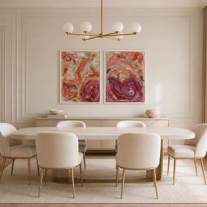 May include: A dining room set featuring an elongated light-colored table with six upholstered chairs on gold-toned metal legs. Above the table, two abstract paintings with red, pink, and orange brushstrokes hang near a gold six-globe chandelier.
