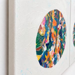 May include: Two square canvases featuring a white textured background with a central circular motif. The circles are filled with thick, impasto-style paint strokes in vibrant shades of orange, blue, green, yellow, pink, and dark brown.