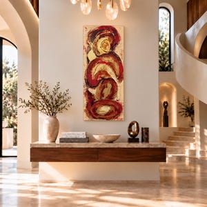 May include: A vertical abstract painting with swirling red, gold, and cream tones hangs above a floating wooden console table. The table holds a ceramic vase with greenery, stacked books, a shallow bowl, and a circular metal sculpture.
