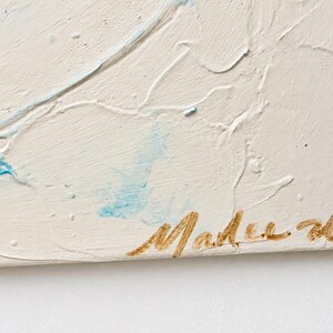 May include: A textured off-white canvas painting featuring raised white paint strokes and subtle light blue accents. The bottom right corner displays a signature in metallic gold cursive script that reads Malee 24.