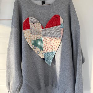 Può includere: Una felpa grigia con un grande cuore patchwork. Il cuore è realizzato con vari modelli di tessuto, tra cui stampe floreali e geometriche in rosso, bianco, blu e verde. La felpa ha un girocollo e maniche lunghe.
