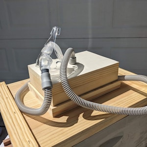 Pode incluir: Uma máscara CPAP transparente com uma mangueira cinza repousa sobre uma caixa de madeira. A mangueira ondulada curva-se em torno da caixa, que fica sobre uma estrutura de madeira maior. A imagem é tirada ao ar livre com luz natural.