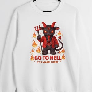 Puede incluir: Sudadera blanca con un diablo de dibujos animados con un tridente, con una chaqueta roja y rodeado de llamas. El texto "GO TO HELL" y "IT'S WARM THERE" está impreso debajo de la ilustración del diablo.