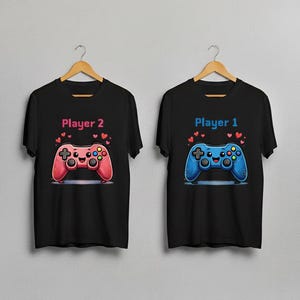 May include: Two black t-shirts with matching video game controller graphics. One shirt says "Player 1" with a blue controller, and the other says "Player 2" with a pink controller. Both controllers have heart accents.