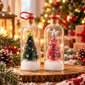 May include: Two clear glass bottle ornaments with cork stoppers and red ribbons. One contains a green bottle-brush tree with a gold star and colorful baubles, the other a pink bottle-brush tree with a silver star and baubles, both on snow.