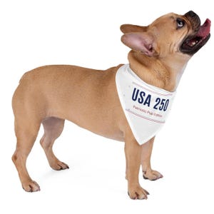May include: A fawn-colored French Bulldog wearing a white triangular pet bandana. The bandana features the dark blue text USA 250 above the red text Patriotic Pup Edition, framed by thin red and blue horizontal lines on the top and bottom edges.