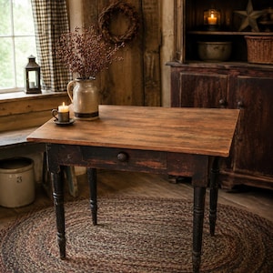 May include: A rectangular wooden table with a natural brown top and a distressed black base featuring a single drawer with a round knob. A ceramic pitcher filled with dried berries and a small mug with a lit candle sit on the table surface.