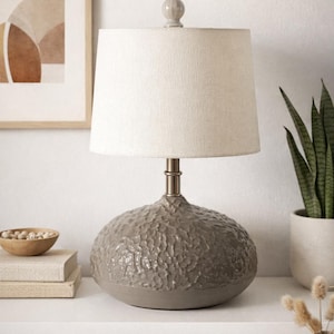 May include: A gray ceramic table lamp with a textured base and a beige fabric shade. The lamp has a silver-colored metal stem and a small, round finial. The lamp is on a white surface, with a framed artwork and a potted plant in the background.