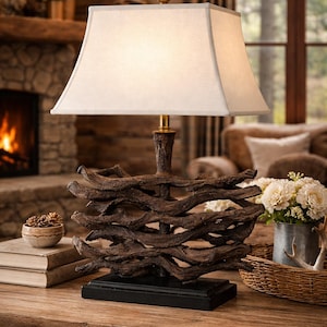 May include: A rustic table lamp with a unique base made of intertwined, dark brown wooden branches. The lamp has a square, off-white lampshade and a brass-colored neck. The lamp sits on a black base, and the overall design evokes a natural, woodland theme.