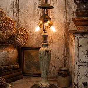May include: A table lamp with a distressed, pale green and brown finish, featuring a central column, a round base, and two illuminated light bulbs. The lamp includes a metal pull chain for operation and a decorative finial at the top.