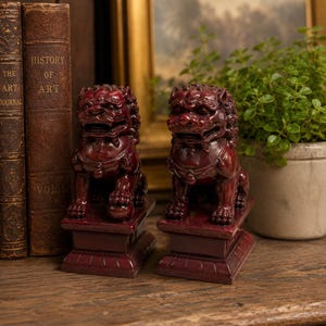 May include: A pair of deep red, glossy ceramic Foo Dog statues, each mounted on a rectangular base. The figures feature intricate manes, open mouths with visible teeth, and detailed collars. One statue rests a paw on a sphere, while the other does not.