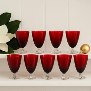 May include: A set of eight ruby red wine glasses with clear, textured bases. The glasses are arranged on a white shelf, with a gold ornament and a white flower with green leaves in the background. The glasses are elegant and suitable for special occasions.