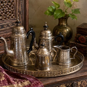 May include: A silver-toned tea set consisting of a tall teapot, a smaller teapot, a lidded sugar bowl, and a creamer pitcher, all arranged on a round, ornate brass tray with a perforated edge and a textured, weathered metallic finish.