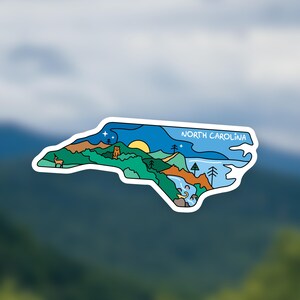May include: A sticker in the shape of North Carolina, featuring a landscape design. The design includes a sun, mountains, trees, a deer, and the text "NORTH CAROLINA" in white. The sticker has a white border.