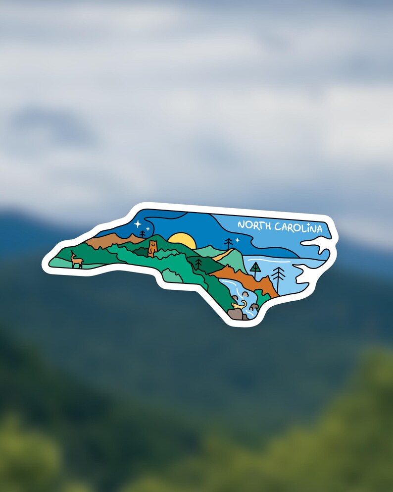 May include: A sticker in the shape of North Carolina, featuring a landscape design. The design includes a sun, mountains, trees, a deer, and the text "NORTH CAROLINA" in white. The sticker has a white border.