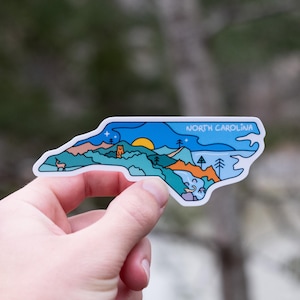May include: A sticker in the shape of North Carolina, featuring a landscape design with mountains, trees, and a sunset. The sticker has the words "NORTH CAROLINA" in a blue banner across the top. The sticker is held in a hand.