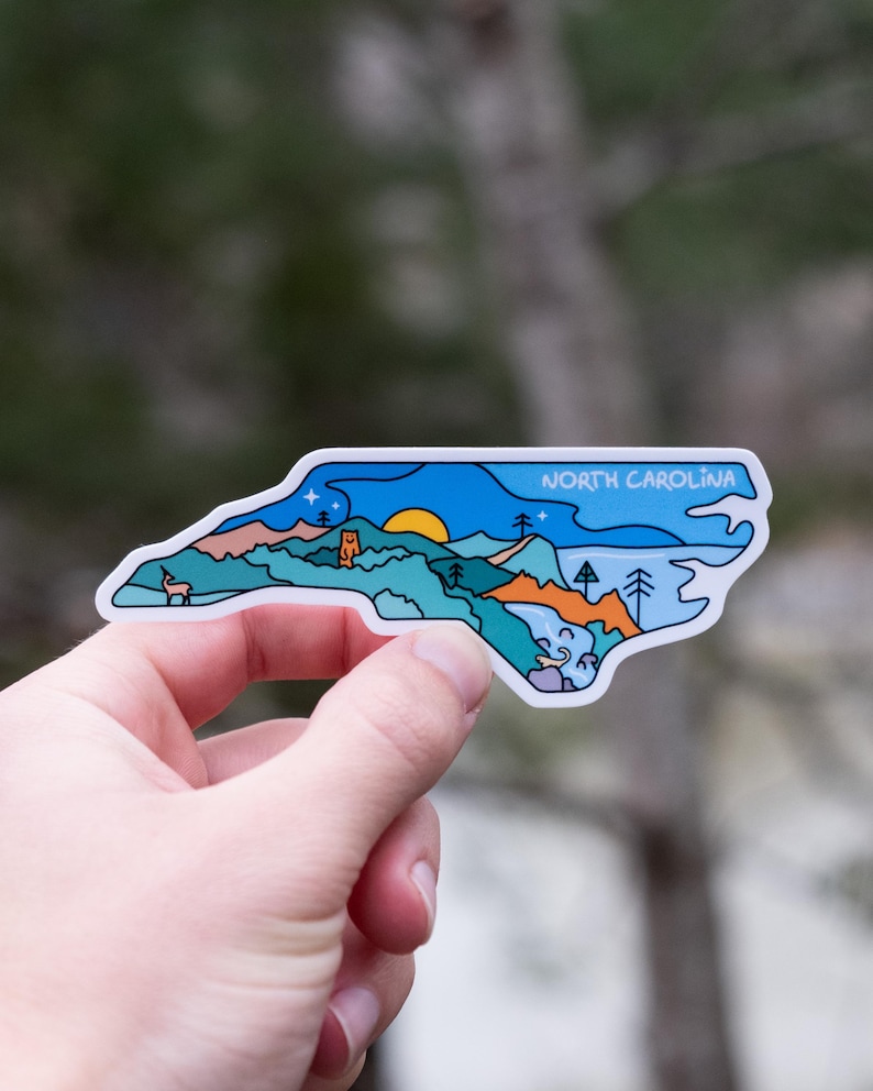 May include: A sticker in the shape of North Carolina, featuring a landscape design with mountains, trees, and a sunset. The sticker has the words "NORTH CAROLINA" in a blue banner across the top. The sticker is held in a hand.